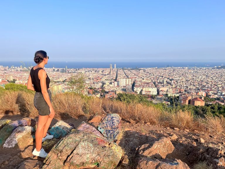 Katharina standing on the rocky viewpoint at Mirador de la Farma, one of the best free things to do in Barcelona, looking out over the panoramic city skyline and the Mediterranean Sea.