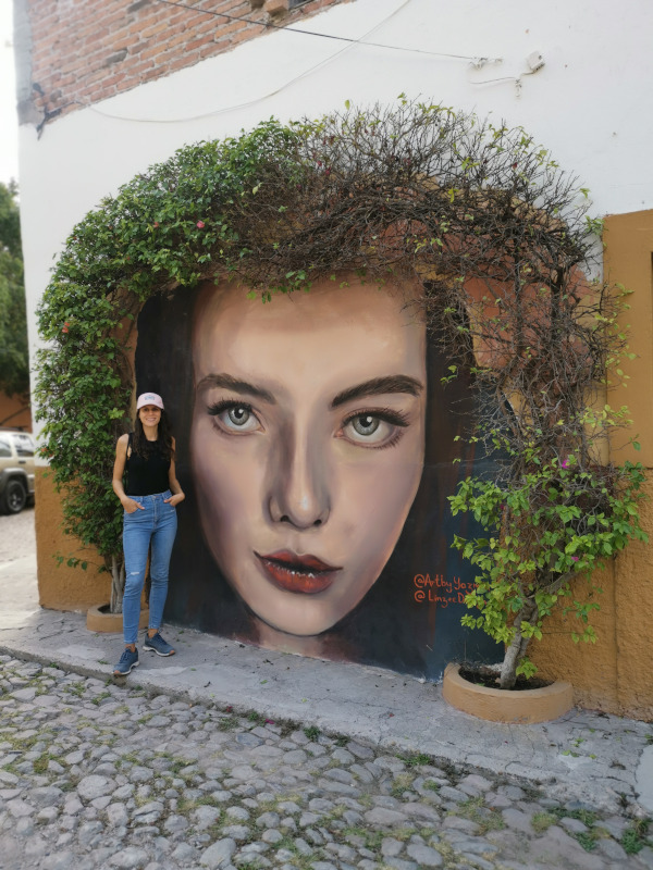 Katharina standing next to an unusual street art of a womans face surrounded by plants as hair in Guadalupe
