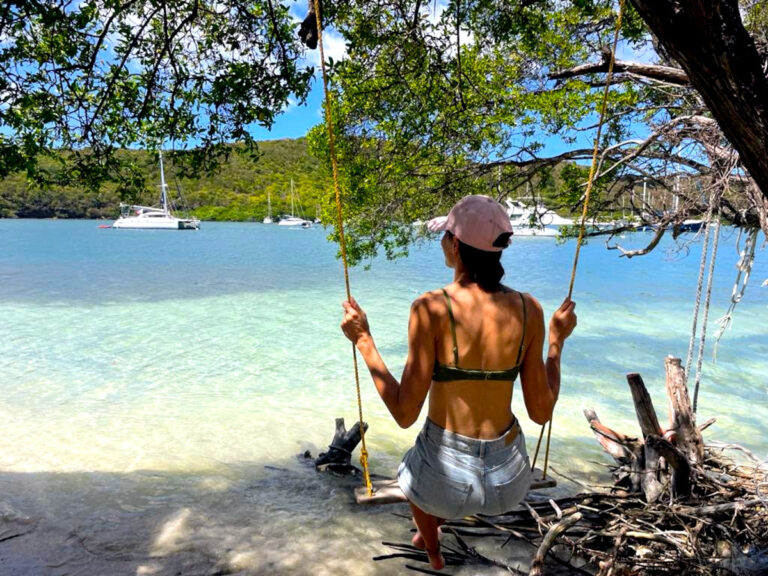 A woman in a bikini top and denim jeans sitting on a swing overlooking clear blue water on hog island grenada