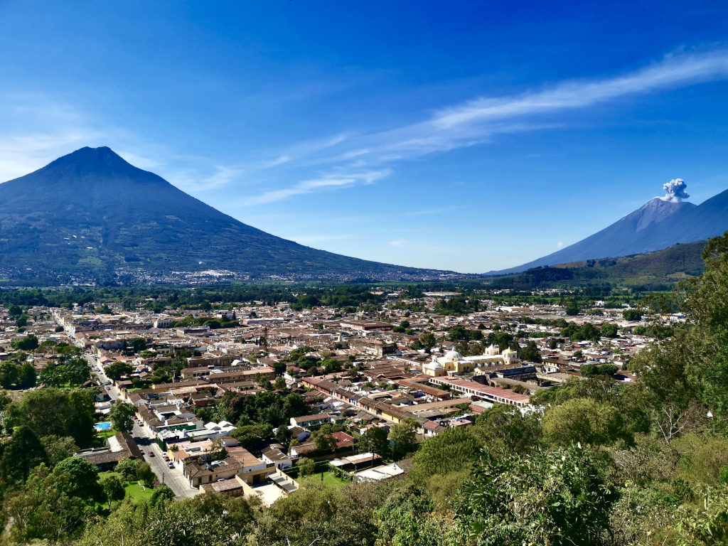 A photo taken of a lookout overlooking the town of Antigua Guatemala with two volcanoes in the background behind town with one of the volcanoes erupting