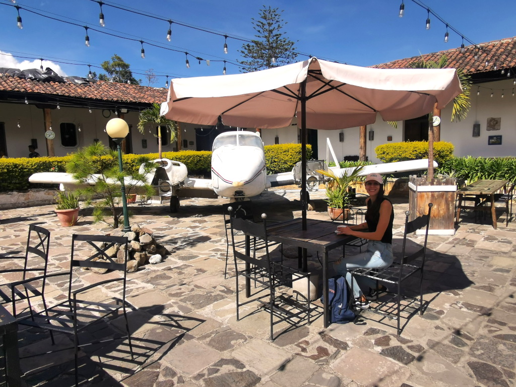 A woman sitting under an umbrella at a cafe in Antigua Guatemala with a coffee cup in her hands and a small airplane in the background
