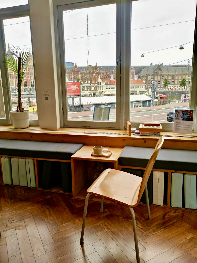 A cup of black coffee on a wooden bench next to a wooden chair in front of a big window overlooking Amsterdam Central Station.