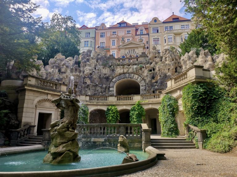 Fountain with a grotto and colorful buildings in the background at Grebovka Park, Vinohrady, Prague.