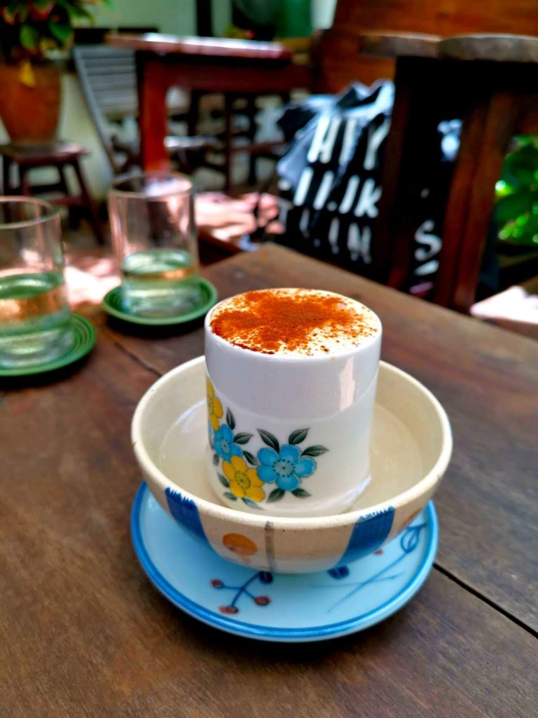 A white floral coffee cup with yellow and blue flowers on it sitting in a small bowl of water with a frothy topping covered in cinammon on a wooden table at a cafe in Vietnam
