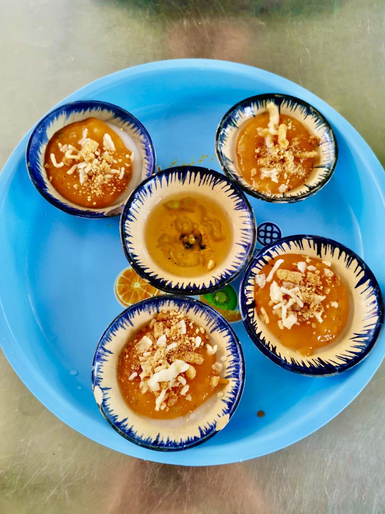 A blue plastic plate with four small bowls of banh beo a traditional vietnamese snack on top. The banh beo consists of a small steamed rice cake topped with savory shrimp, crispy pork bits and a bowl of fish sauce in the middle to drizzle over the cakes.