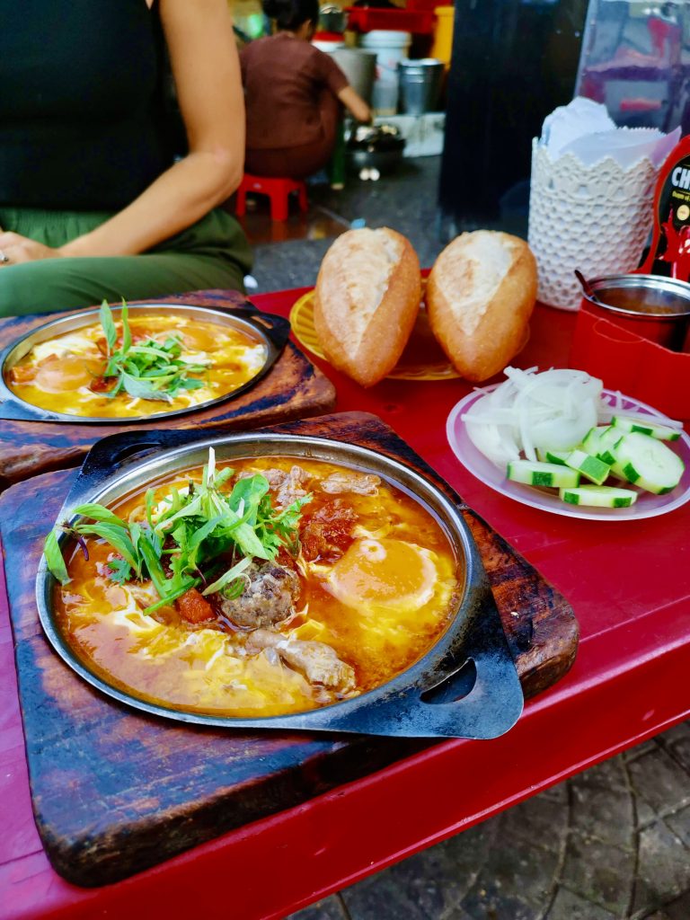 A classic Hoi An food breakfast featuring sizzling pans of bánh mì ốp la with fried eggs, meatballs, and herbs in rich tomato sauce, served with crispy baguettes, sliced cucumber, and onions at a red street-side table.