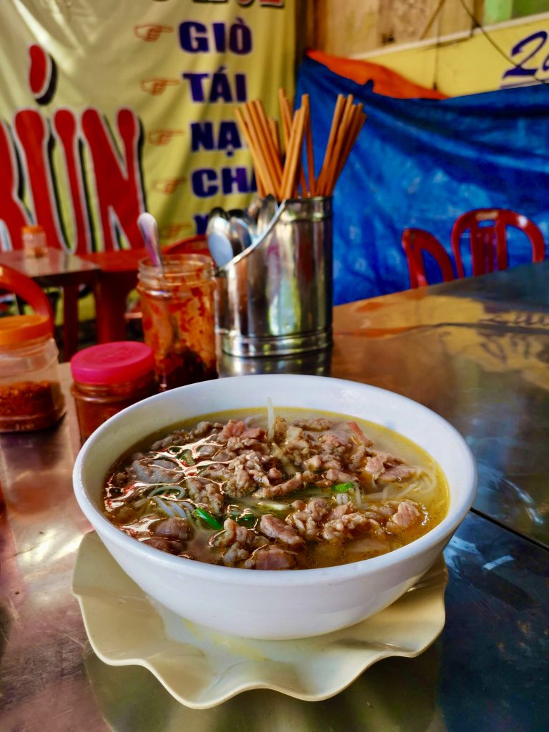 A white bowl of bun bo a meaty and spicy soup sitting on top of a metallic table at a street food stand in hoi an, behind the bowl are containers of hot sauce choptsticks and spoons and red plastic chairs for seating at the table.