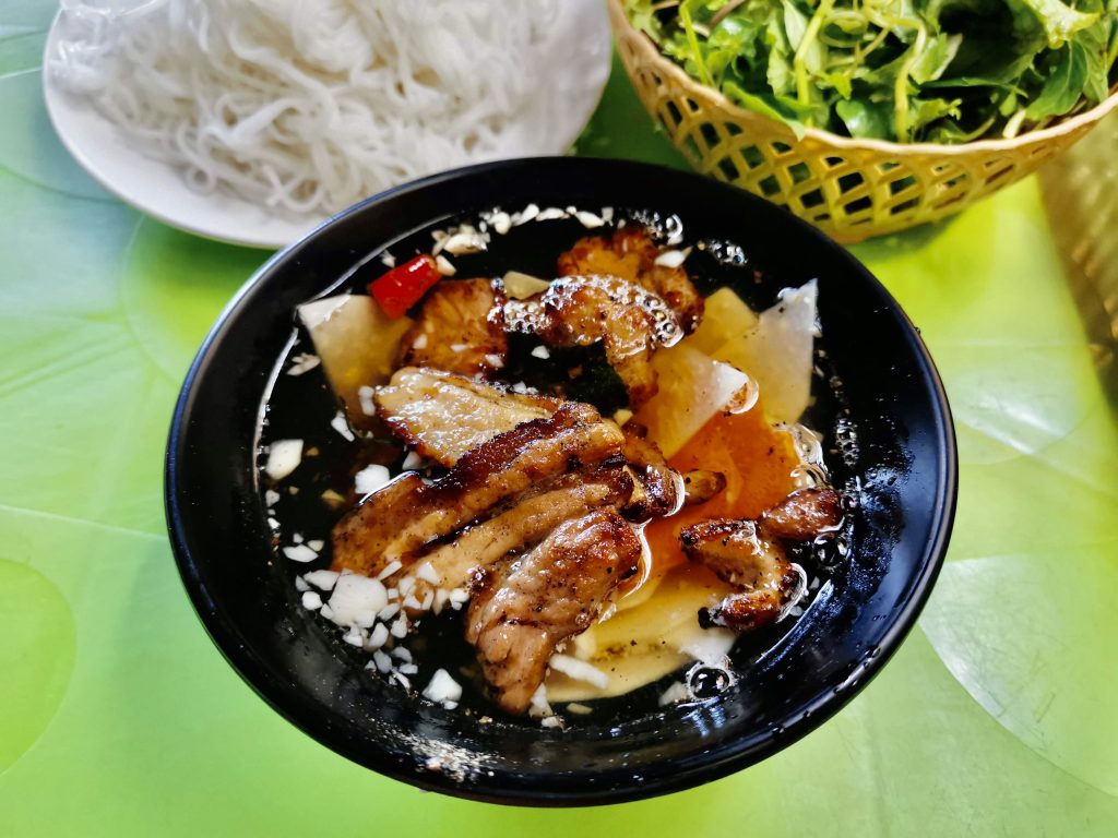 A black bowl of Bun Cha containing freshly grilled pork in a sweet sauce with grilled carrot and onion. Behind the meat is a white plate with noodles and a basket of fresh green herbs.