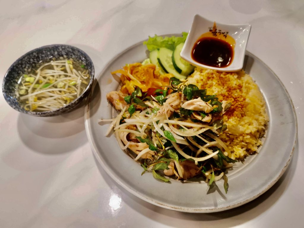 A plate of cơm gà, featuring shredded chicken with fresh herbs and bean sprouts, turmeric rice, pickled vegetables, cucumber slices, and soy sauce, served with a side bowl of clear broth with bean sprouts.