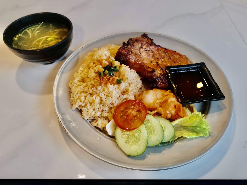 A savory plate of cơm tấm, a popular dish, with grilled pork chop, broken rice topped with scallion oil, pickled vegetables, lettuce, tomato, cucumber slices, and a side of dipping sauce, served with a bowl of clear broth.