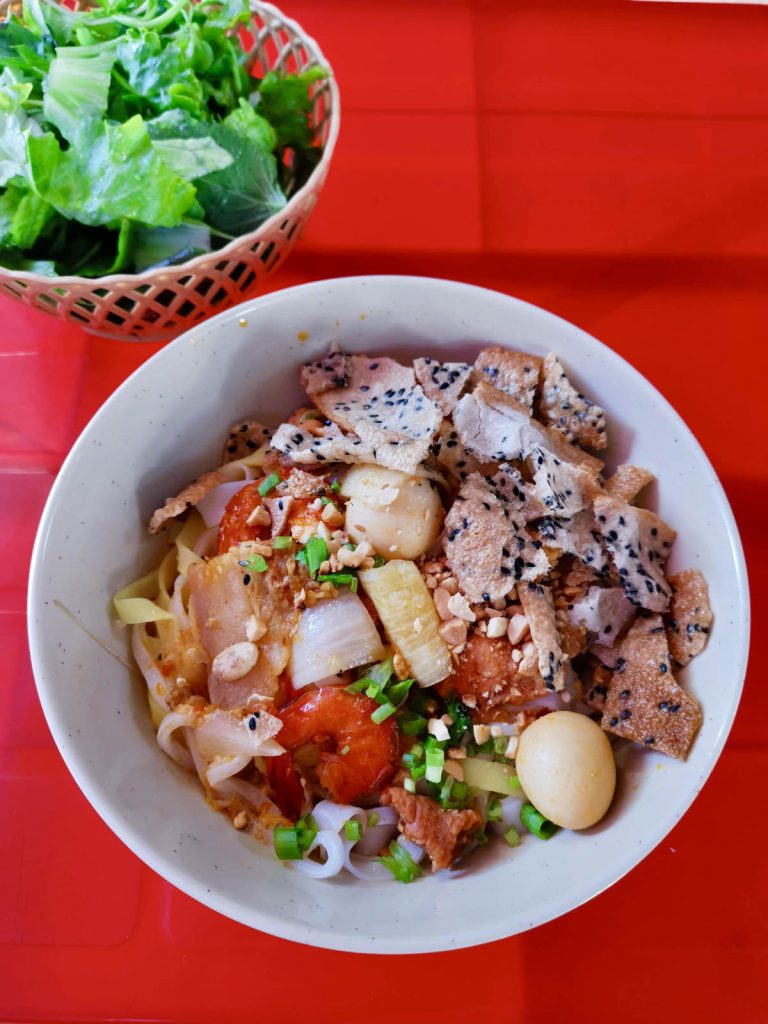 A vibrant bowl of mì Quảng, a signature Hoi An food, featuring turmeric rice noodles topped with shrimp, pork, quail egg, crushed peanuts, scallions, sesame rice crackers, and diced vegetables, served alongside a basket of fresh herbs on a red table.