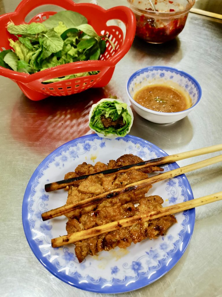 A serving of Thit Nuong Xien a dish consisting of skewers of freshly grilled succulent pork, a bowl of peanut sauce and freshly picked local herbs sitting on top of a shiny metallic table.