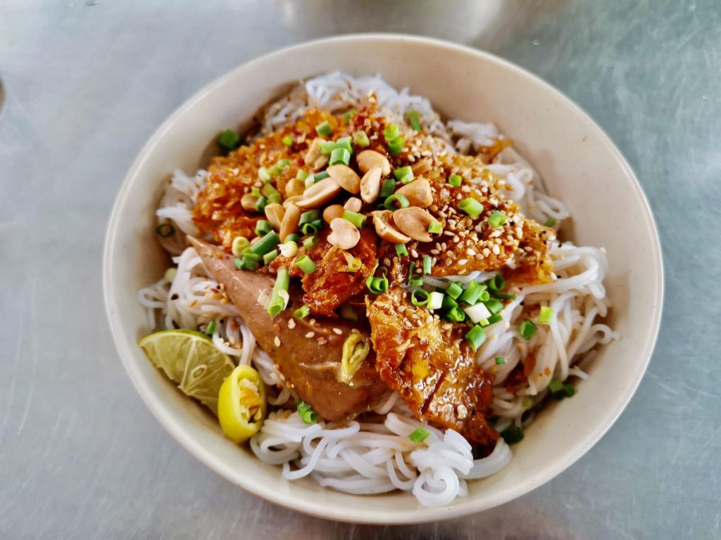 A bowl of bún thịt nướng, a popular dish made with rice vermicelli noodles, grilled pork, fried shallots, chopped scallions, crushed peanuts, and a slice of lime, all served on a metallic tabletop.