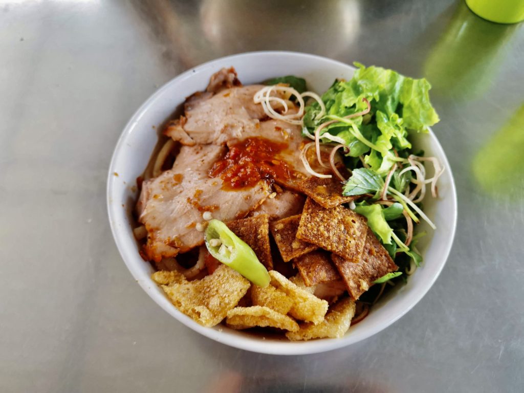 A bowl of Hoi An food specialty, cao lầu, served with thick rice noodles, slices of roast pork, crispy fried wonton pieces, fresh herbs, lettuce, a green chili pepper, and spicy red chili sauce, all presented on a shiny metal table.