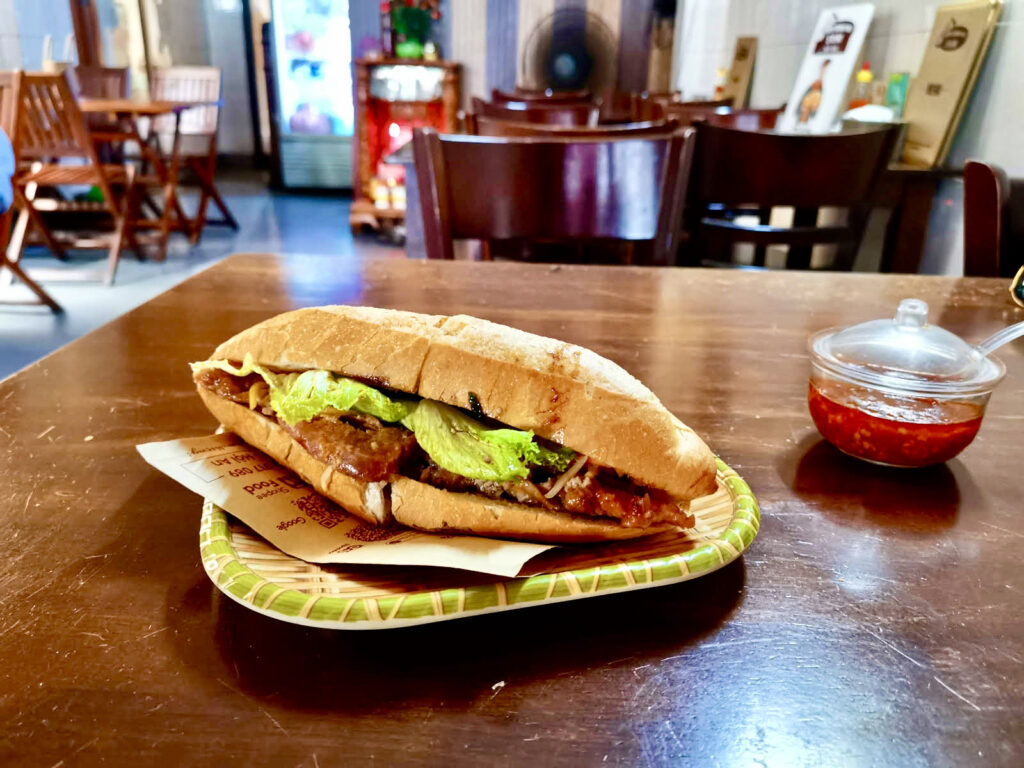 A fresh bánh mì sandwich, a beloved Vietnamese food, filled with grilled meat, pickled vegetables, and lettuce in a crispy baguette, served on a bamboo-patterned plate in a cozy Vietnamese eatery with a jar of red chili sauce on the table.