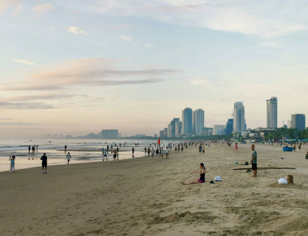Locals and visitors stroll and relax along Đà Nẵng’s beach at sunrise, with soft pink skies over high-rise buildings and a gently receding tide creating reflections on the wet sand.