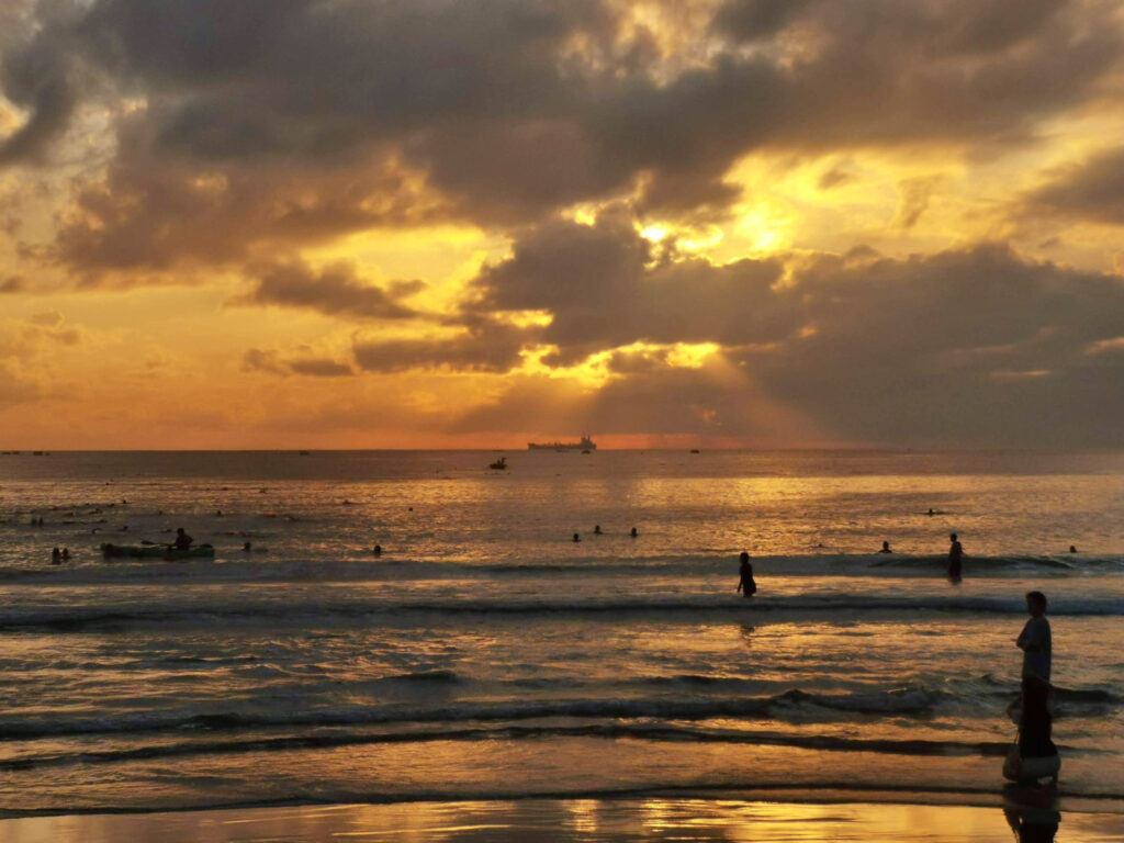 Locals swim in the water at Da Nang during an orange sunrise as a container ship sits in the water on the horizon underneath the sun