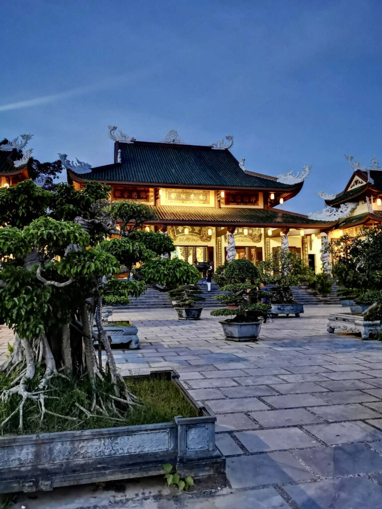 A serene evening view of a beautifully lit temple near the Lady Buddha statue in Da Nang, with bonsai trees in stone planters lining the tiled courtyard. The ornate roof and dragon carvings stand out against the deep blue sky, reflecting Da Nang’s peaceful yet grand spiritual atmosphere.
