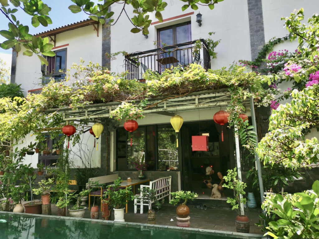 A man crouching down to pat a white dog in the doorway of a house that is surrounded by green foliage and colorful lanterns with a pool in the foreground