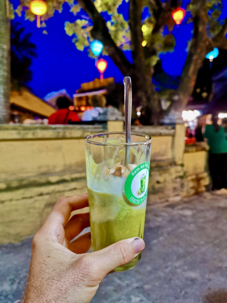 A hand holds a tall glass of Vietnamese avocado ice cream with coconut, partially melted, in front of a lantern-lit evening street scene in Hoi An.