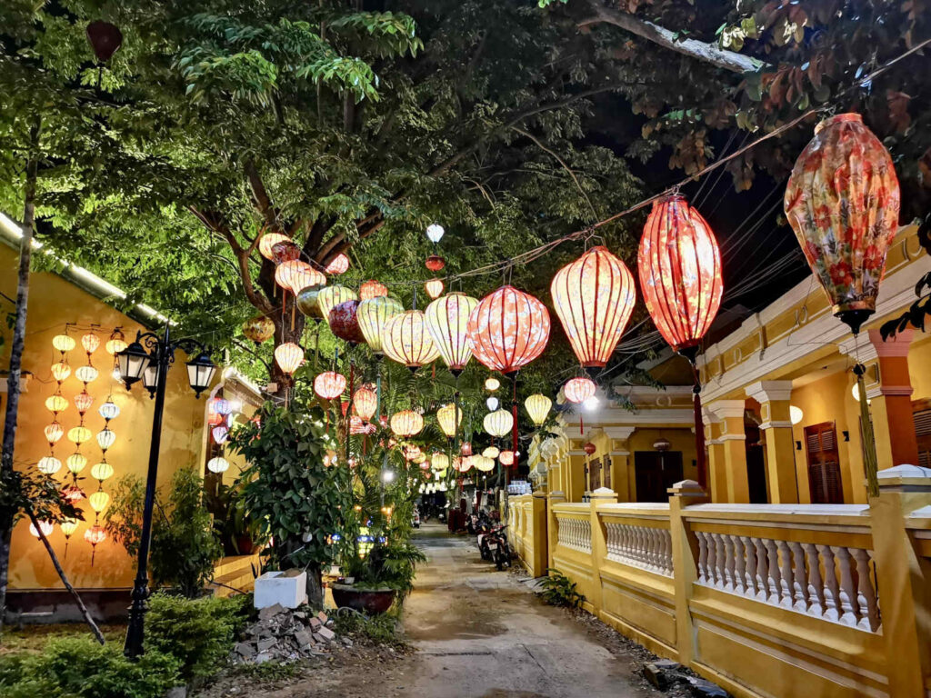 A small alleyway in Hoi An at night time with colorful lanterns strong up across the alley and yellow and white colonial buildings lining the alley