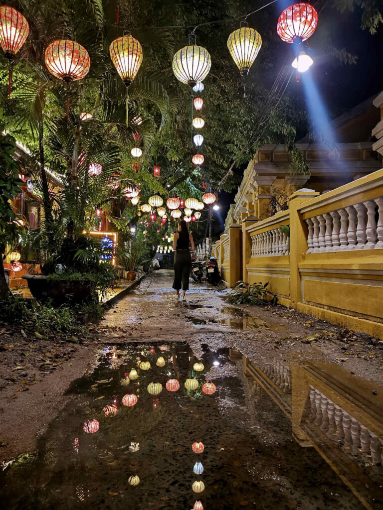 A narrow, lantern-lit street in Hoi An at night, with colorful hanging lanterns reflecting in a large puddle on the ground. A woman walks down the path between lush greenery and yellow colonial-style buildings, capturing the old-world charm that defines Hoi An.
