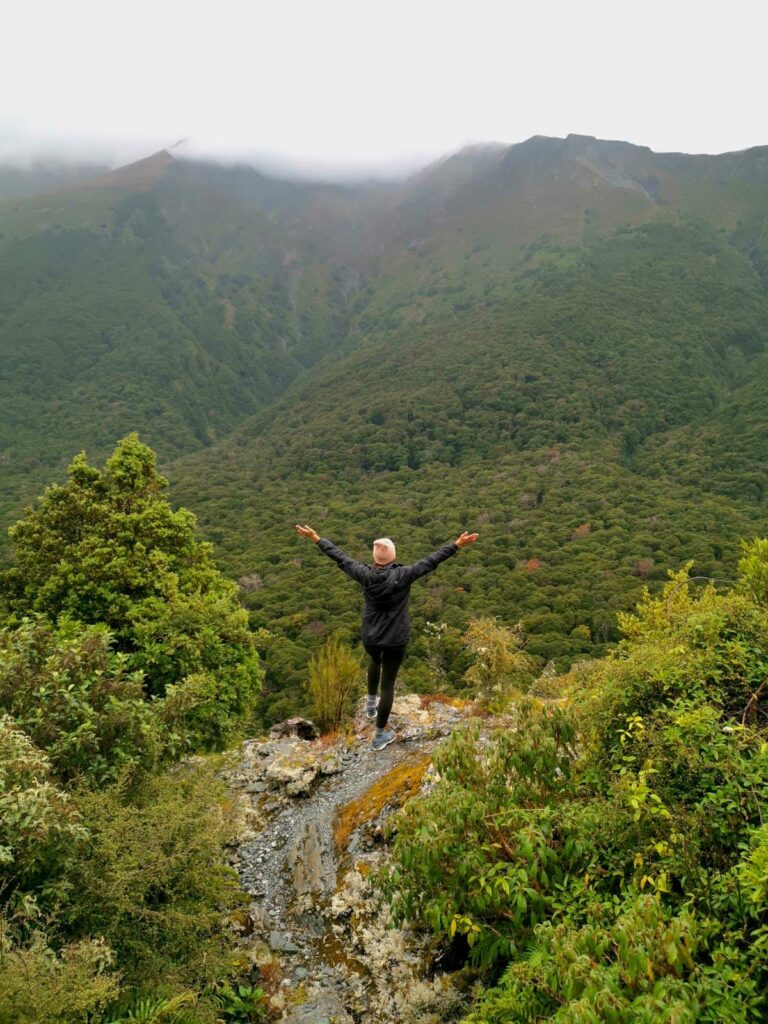 Katharina standing with arms raised on a rocky forest trail in Haast Pass, surrounded by lush green mountains in New Zealand under a misty sky.