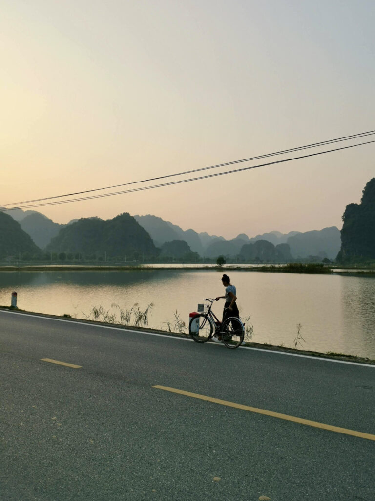 Katharina standing with a bicycle on the side of a road, looking at a calm body of water with misty karst mountains in the background at sunset in Ninh Binh, Vietnam.