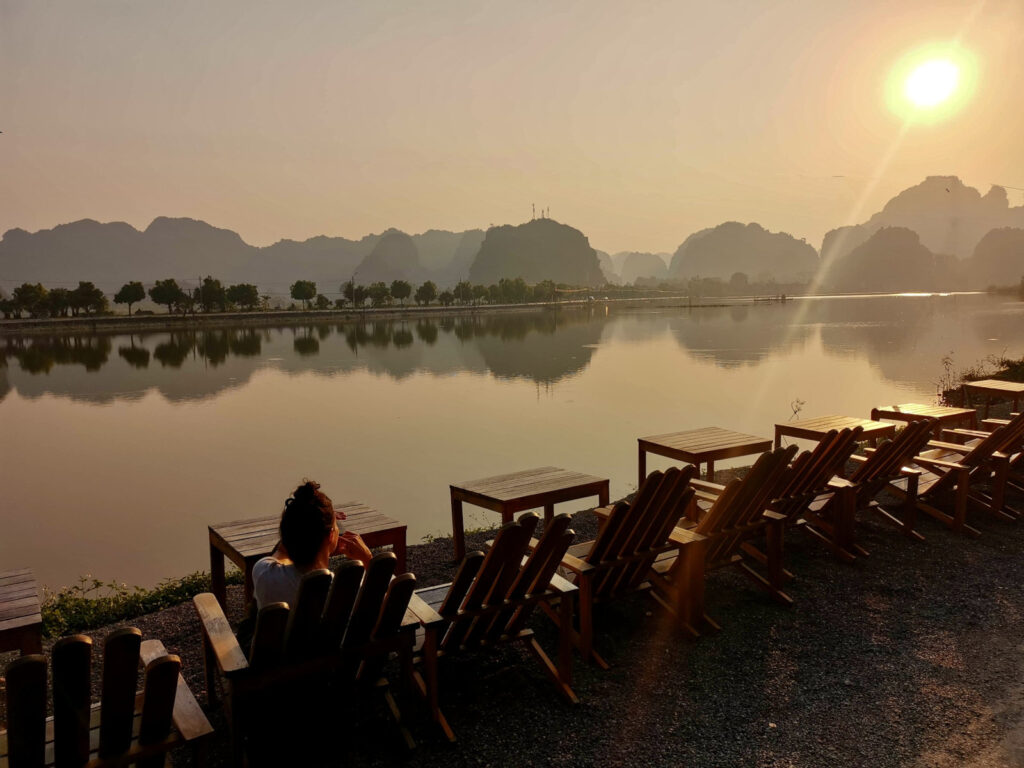 Katharina sitting at a cafe table, enjoying the sunset over a calm lake and the silhouette of the karst mountains in Ninh Binh, Vietnam.