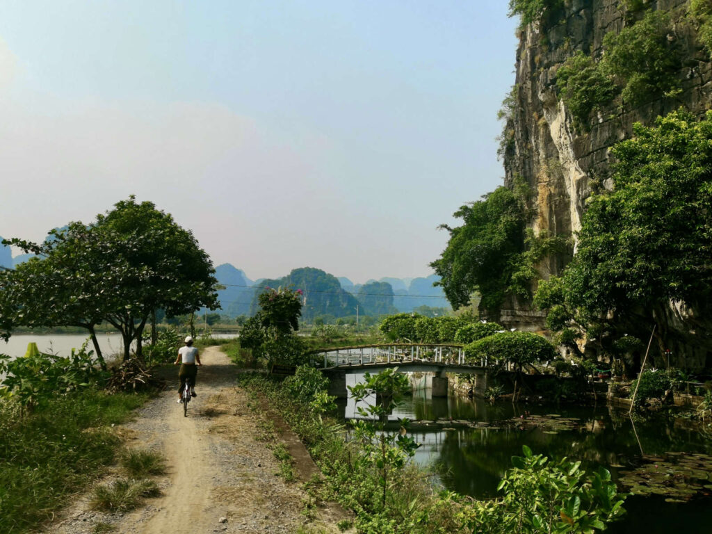 Katharina riding a bicycle along a dirt path next to a river, with a small bridge and lush green karst mountains in the background of Tam Coc, Ninh Binh.