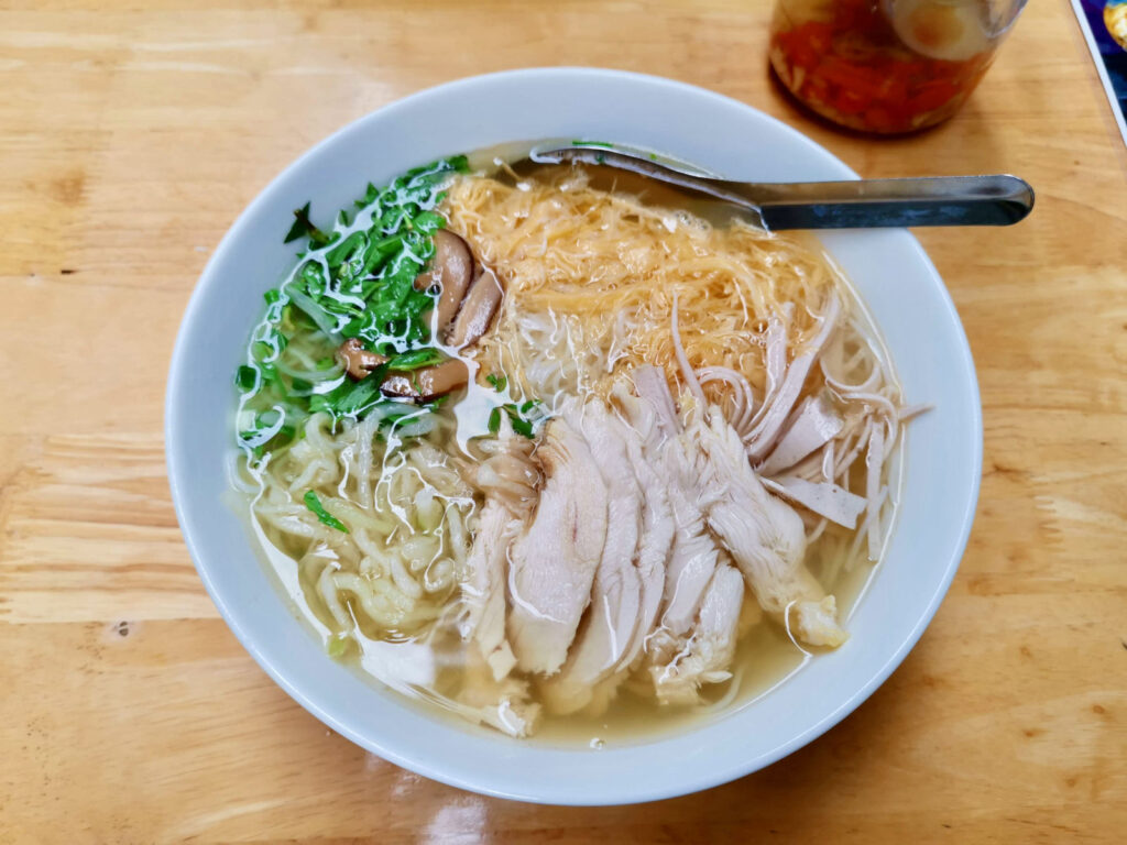A close-up, top-down view of a bowl of traditional Vietnamese chicken pho. The dish features a rich, clear broth with rice noodles, sliced chicken, fresh green herbs, and a garnish of red chili slices.