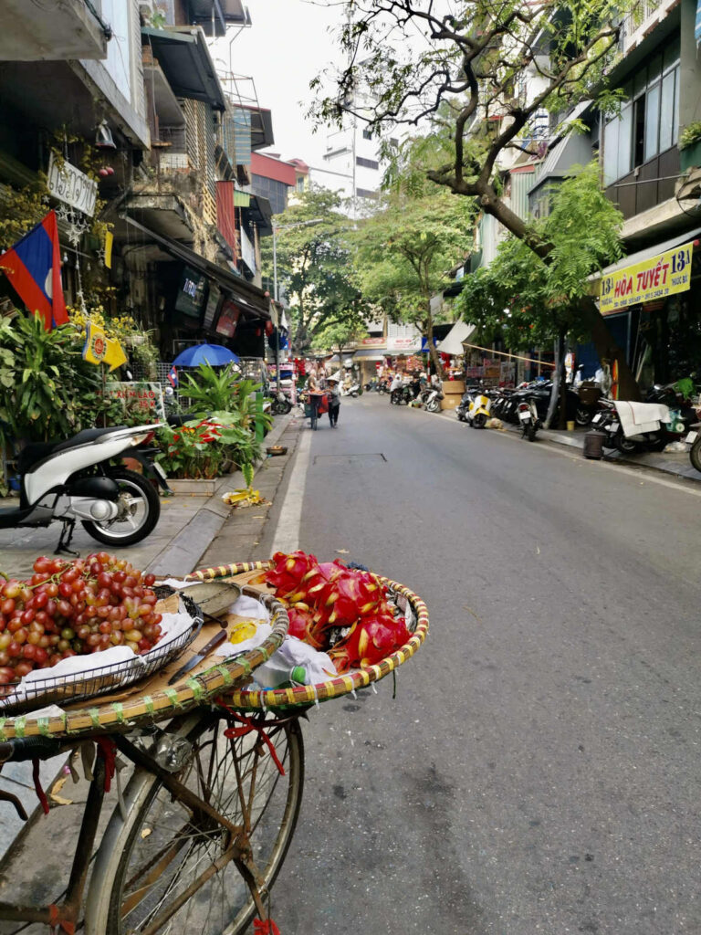 A close-up of a bicycle street vendor's cart, laden with baskets of fresh red grapes and vibrant red dragon fruit. In the background, a busy Hanoi street is visible with motorbikes parked along the curb, and people walking on the sidewalk.
