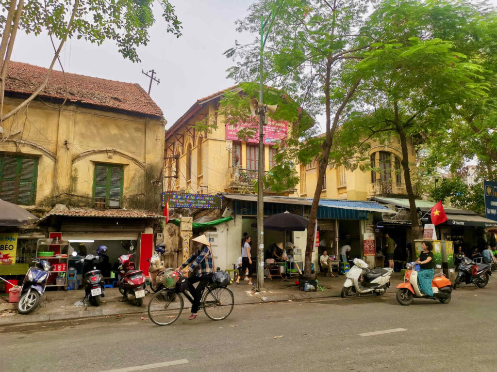 A vibrant street scene in Hanoi, with a person riding a bicycle past old buildings with faded yellow and brown facades. Motorbikes are parked along the sidewalk, and leafy trees overhang the street, capturing the typical daily life in the city.
