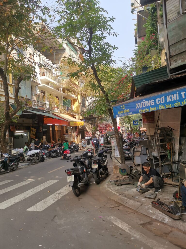 A bustling street in Hanoi with motorbikes parked on the left side of the road. On the right, a person is seen working on metal parts outside a workshop, surrounded by tools and equipment. The buildings are multi-story with a mix of commercial and residential use.