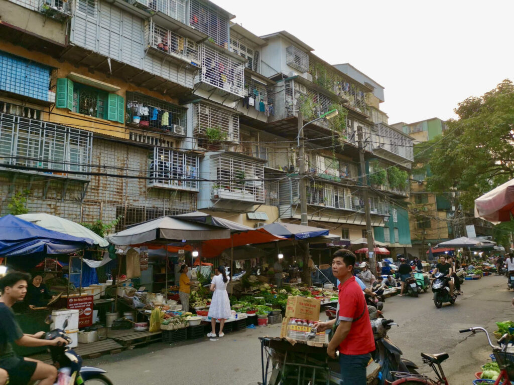 A busy Hanoi street scene with market stalls under large awnings. People are shopping for produce and other goods. In the background, tall, multi-story residential buildings with balconies and grates rise up. Motorbikes are parked and a person wearing a red shirt stands next to a cart in the foreground.