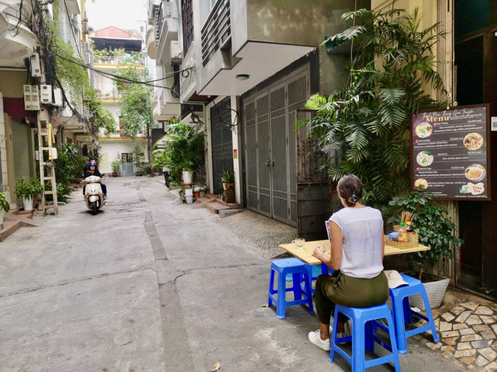 A close-up view of Katharina eating at a small, low table set up on the sidewalk in a Hanoi alley. A handwritten menu board hangs next to the table, while a person on a motorbike drives past in the distance. This image captures the intimate atmosphere of local street food culture.