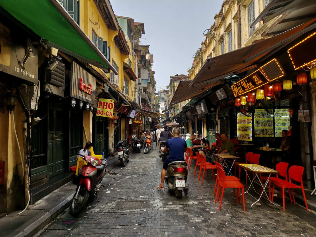 A cobblestone street in Hanoi, packed with motorbikes and people. On the right, a row of restaurants with red plastic chairs and tables spills onto the sidewalk. The buildings are brightly lit with signs and lanterns, capturing the lively evening atmosphere