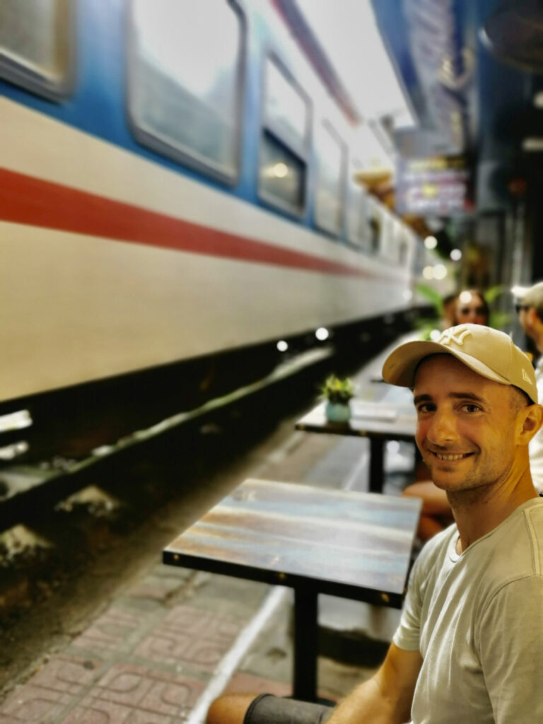 A man in a baseball cap smiles at the camera while sitting at a small, rustic cafe table. A train passes inches from the table, its blue and white cars blurred by motion, creating a thrilling and intimate moment on the Hanoi Train Street.