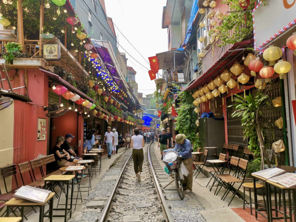 A vibrant daylight scene on the famous Hanoi Train Street, with railway tracks running down the center of a narrow street. Colorful decorative lanterns and flags hang above the buildings on both sides, which are lined with small wooden cafe tables and chairs. People are walking on and alongside the tracks.