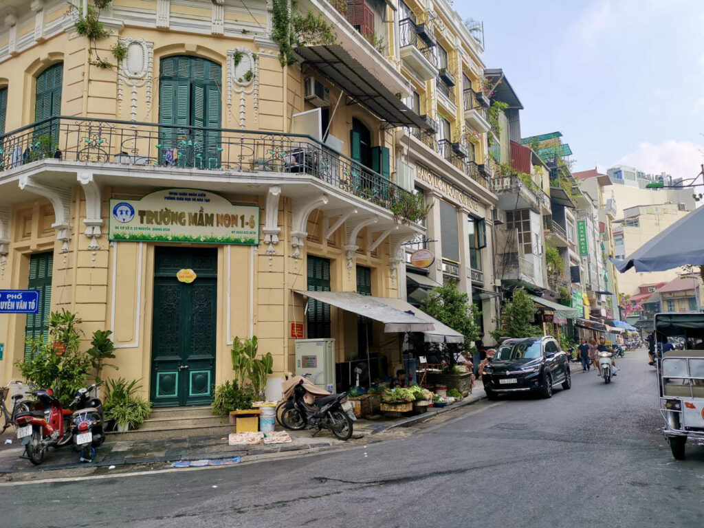 A view of a yellow-painted building with arched windows and green shutters, featuring a sign for a kindergarten. The building is on a busy street with motorbikes parked on the side. In front, a small vendor's stand with fresh produce is set up, capturing a mix of historic architecture and modern daily life.