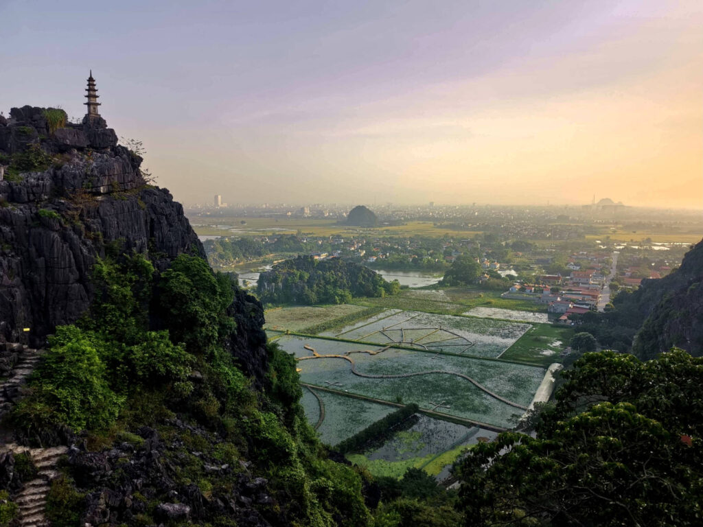 An asian style temple on the pinnacle of a steep limestone cliff overlooking rice fields and a town during sunrise in Ninh Binh Vietnam