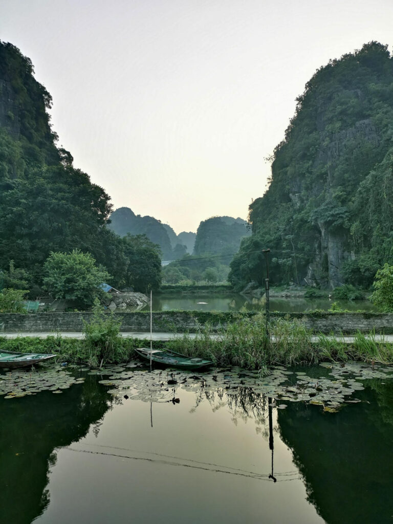 The reflection of towering karst mountains and lush greenery in a calm body of water, with a small boat and water lilies in the foreground of Ninh Binh, Vietnam.