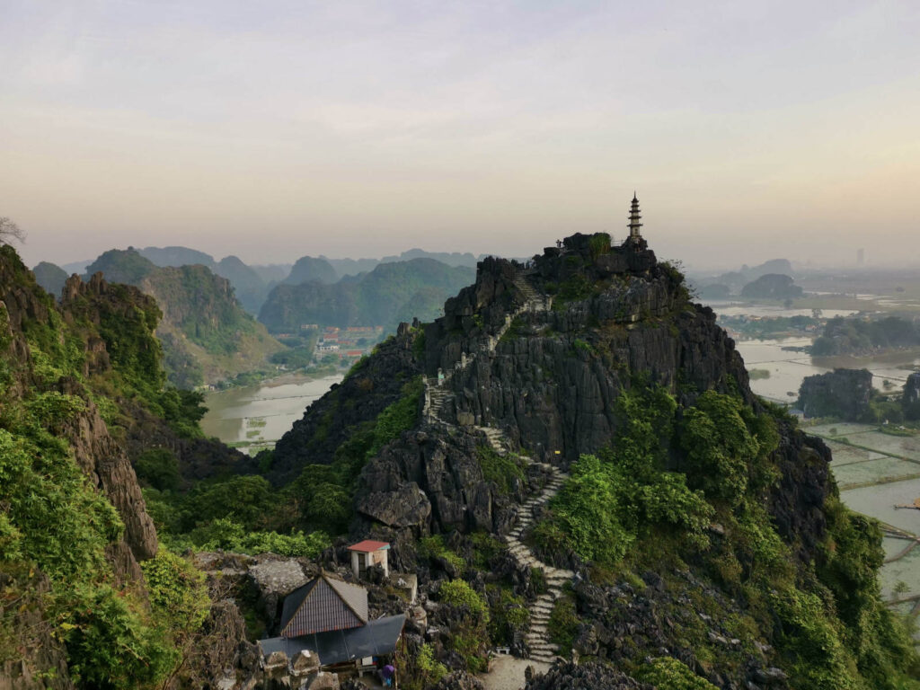 A wide-angle view from the top of Mua Caves, showing the intricate stone staircase leading to a small pagoda on the peak, with a panoramic view of the vast green rice paddies and karst mountains stretching into the misty horizon.