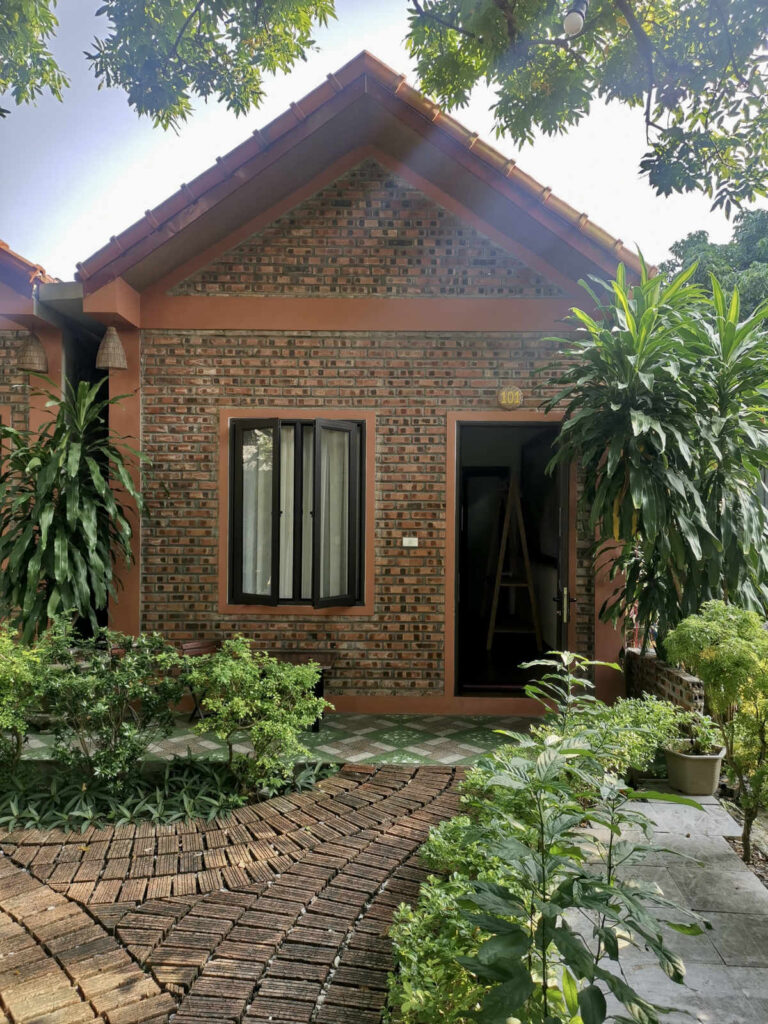 A charming brick bungalow with a tiled roof, surrounded by lush green plants and a pathway leading to the front door, representing a typical homestay in Ninh Binh