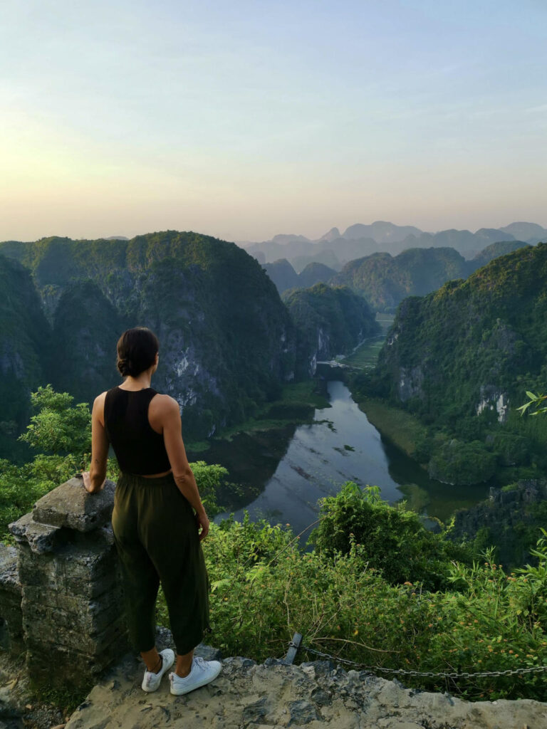 Katharina standing on a stone viewpoint overlooking the lush green limestone karsts and winding river valleys of Ninh Binh, Vietnam, during sunrise.