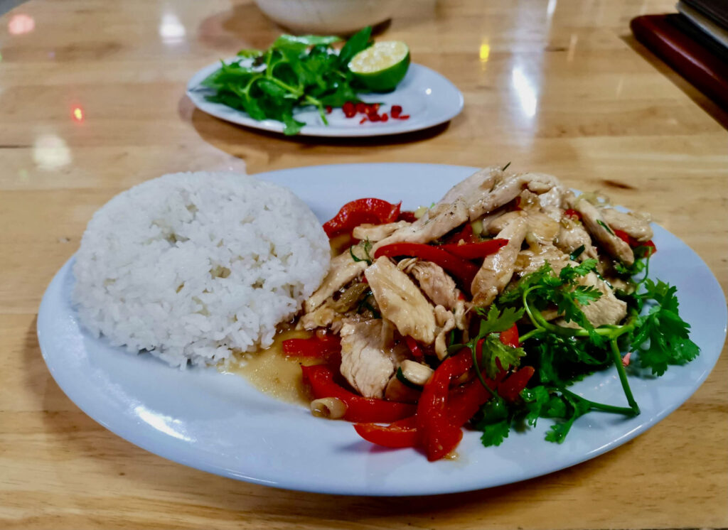 A close-up of a plate of a traditional Vietnamese dish with sliced chicken, red bell peppers, and fresh herbs, served with a side of steamed rice on a wooden table.