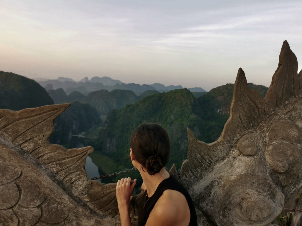 View from the top of Mua Cave viewpoint in Ninh Binh, Vietnam, showing a winding stone dragon sculpture along the ridge at sunrise, with mountains and rice fields in the distance.