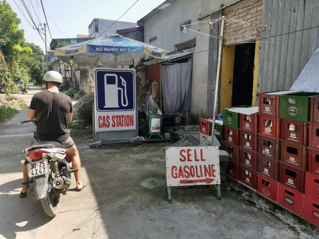 Man sitting on a scooter next to make shift gas station on the messy backstreets of a vietnamese town with piles of plastic crates full of beer bottles next to him