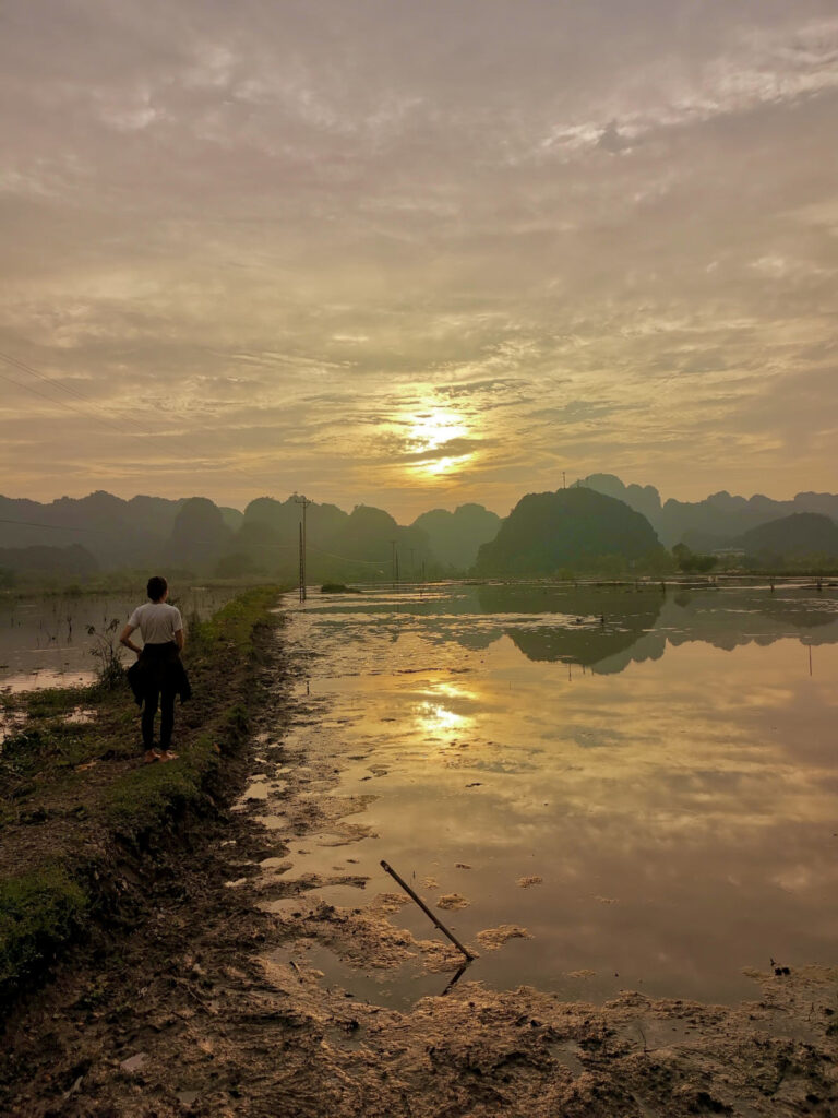 A person stands on a muddy path next to a water-logged rice field, watching the sunset over the distant karst mountains, which are reflected in the still water.