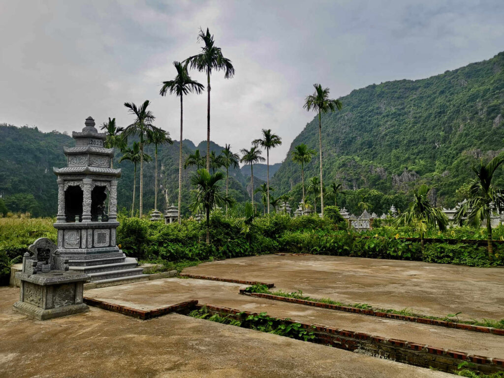 An ornate, carved stone tomb in a Vietnamese cemetery, surrounded by palm trees and lush green karst mountains under a cloudy sky in Ninh Binh.
